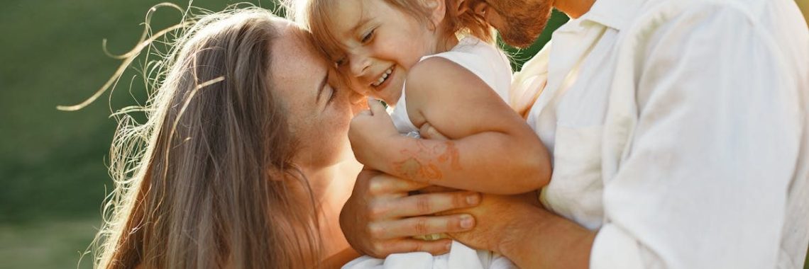 A joyful family moment with parents embracing their child outdoors in warm sunlight.